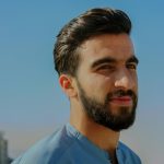 Home A close-up outdoor portrait of a young man with facial hair, exuding confidence under a clear sky.